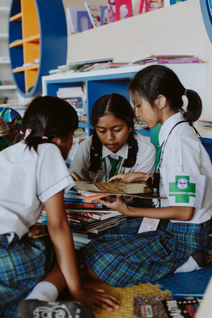 Three girls in school uniforms enjoying reading books together in a colorful library.