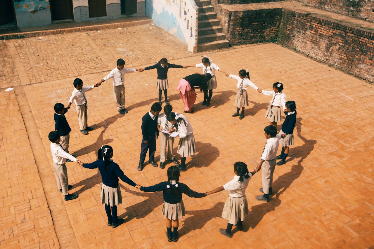 Children in school uniforms playing a circle game outdoors under a teacher's supervision.